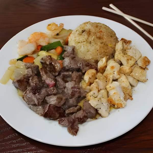 Hibachi meal with steak, chicken, shrimp, fried rice, and vegetables on a white plate with chopsticks.